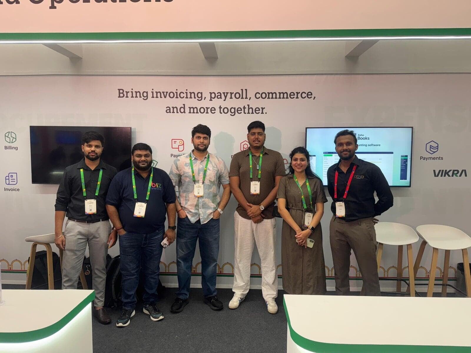 The Biz Systems team with a Zoho team member at the Zoho Finance booth, in front of a wall reading Bring invoicing, payroll, commerce, and more together